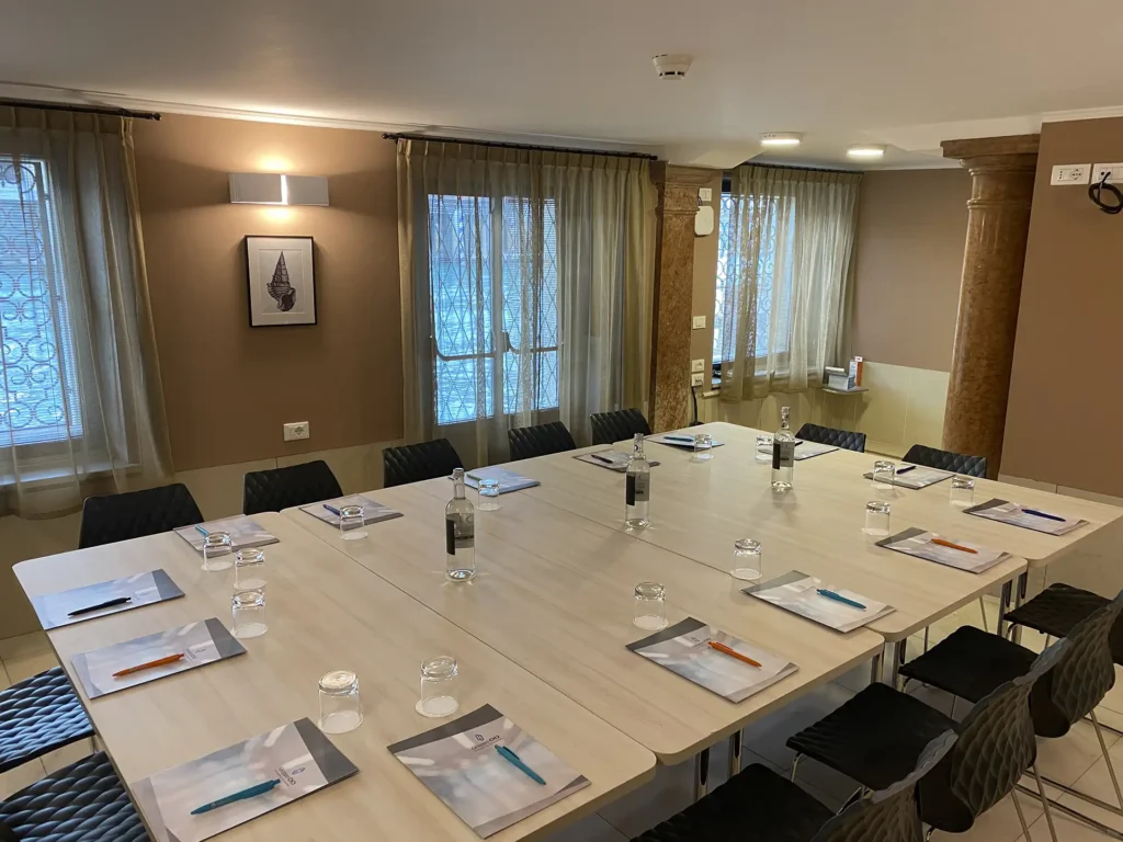 Nell'immagine la sala meeting dell'hotel con al centro un grande tavolo unico di legno chiaro circondato da sedie di plastica nera. Sul tavolo ci sono delle cartelline, penne e bottiglie d'acqua. Dalle finestre sulla parete di sinistra si vede il Canal Grande.