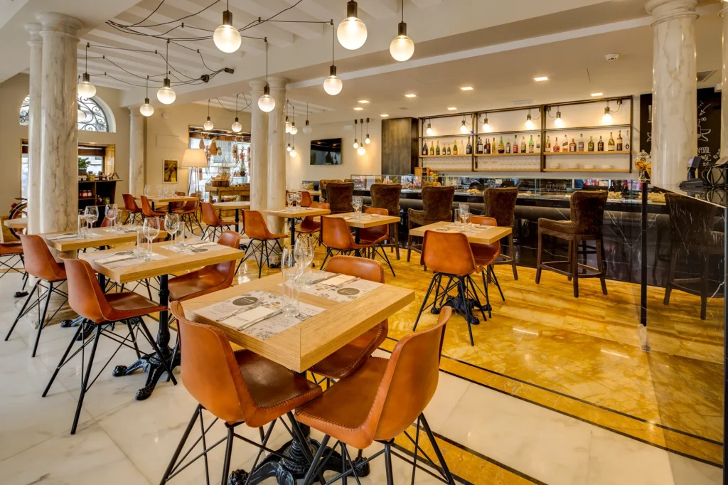 Nell'immagine il Cicchetti Bistrot interno all'hotel. La sala è occupata da tavoli di legno chiaro con gambe in ferro battuto e sedie arancio con gambe nere. Il pavimento è di marmo bianco e giallo. A destra c'è il bar dell'hotel con il bancone, degli sgabelli di velluto e mensole di alcolici. Il soffitto è composto da travi bianche e lampadari bianchi tondi sospesi collegati da fili neri uno all'altro.