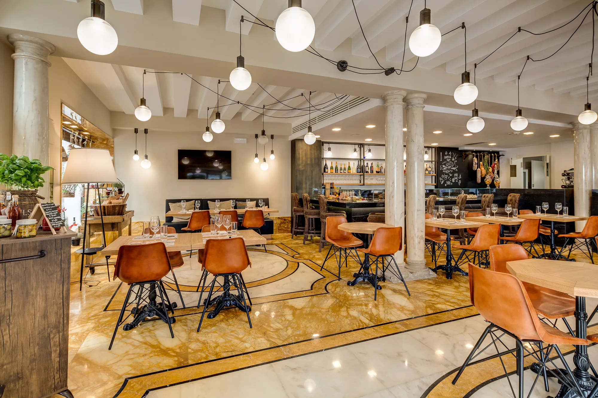 Nell'immagine il Cicchetti Bistrot interno all'hotel. La sala è occupata da tavoli di legno chiaro con gambe in ferro battuto e sedie arancio con gambe nere. Il pavimento è di marmo bianco e giallo. In fondo a destra c'è il bar dell'hotel con il bancone e le mensole di alcolici. Il soffitto è composto da travi bianche e lampadari bianchi tondi sospesi collegati da fili neri uno all'altro.