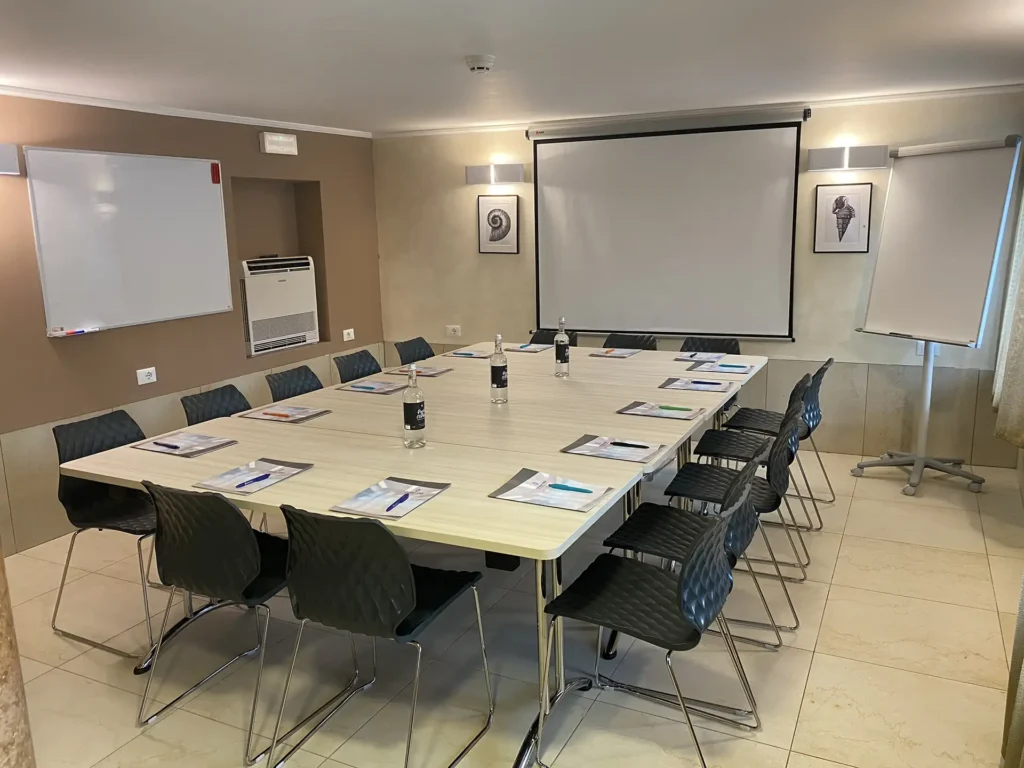 Nell'immagine la sala meeting dell'hotel con al centro un grande tavolo unico di legno chiaro circondato da sedie di plastica nera. Sul tavolo ci sono delle cartelline, penne e bottiglie d'acqua. Sulla parete di fronte c'è un telo per proiezione e, accanto, una lavagna a fogli mobili.