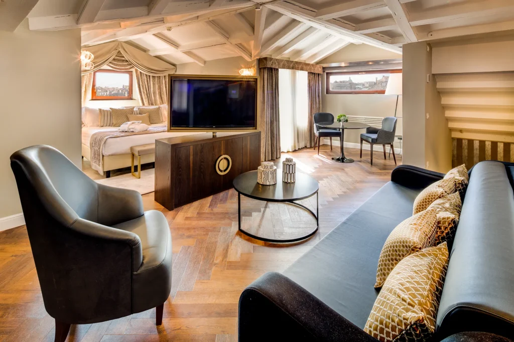 Nell'immagine la suite dell'hotel. In primo piano c'è la zona living con divano di pelle nera e cuscini, poltroncina nera e al centro tavolino basso nero. Sopra un armadietto basso di legno scuro c'è una tv girevole. Dietro la tv si vede il letto matrimoniale sopra il quale c'è una piccola finestra. Sulla destra, in fondo. c'è un tavolino nero e due sedie. Dietro il tavolino si vede una finestra rettangolare orizzontale e a destra auna finestra coperta da tende. Il soffitto è mansardato di color bianco.
