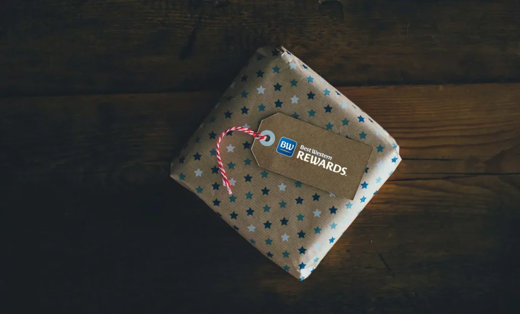Nell'immagine c'è un pacco regalo posato su un piano di legno. Il pacco ha una carta color marrone con delle stelle blue azzurre. Sopra è posato un tag di carta marrone con il logo Best Western Rewards®