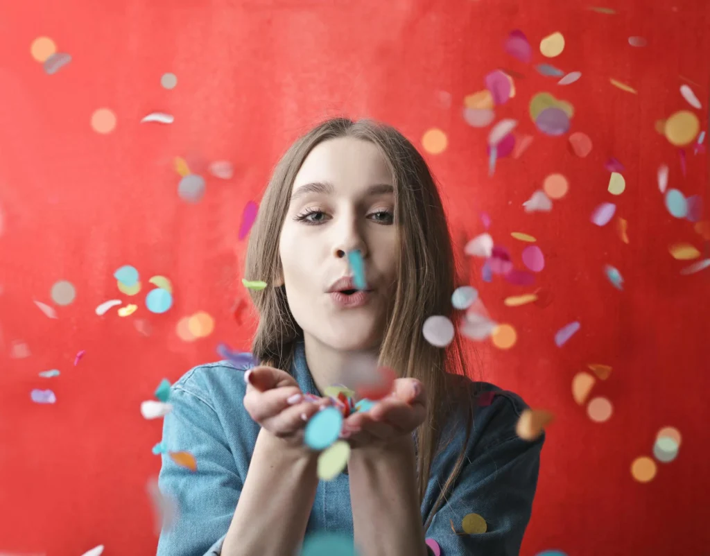 Nell'immagine una ragazza ha le mani aperte davanti a sè con all'interno dei coriandoli colorati che soffia in aria. Lo sfondo dell'immagine è rosso.