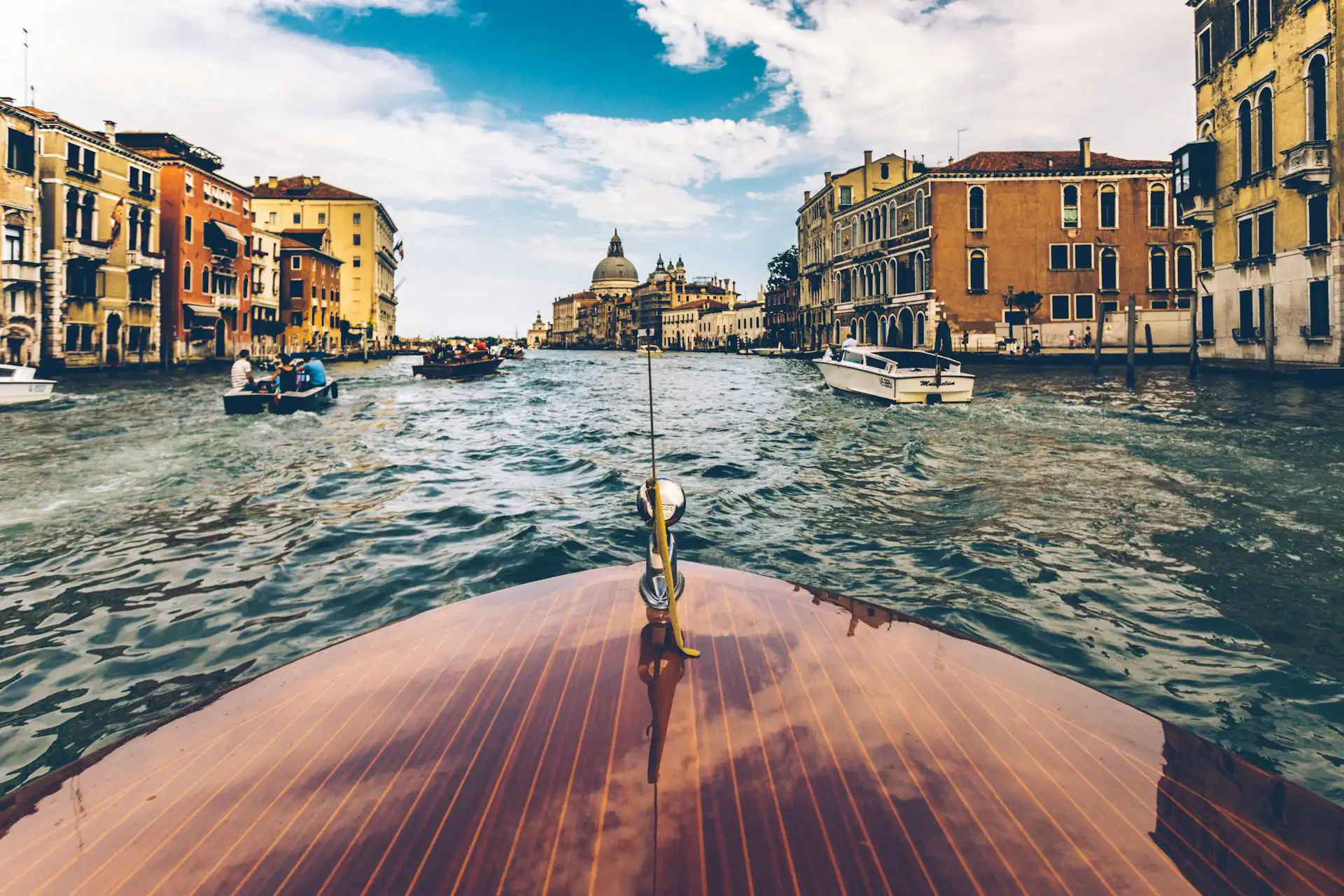 Nell'immagine c'è in primo piano la prua di una barca che naviga in uno dei canali di Venezia. Sui lati ci sono le facciate dei palazzi della città.