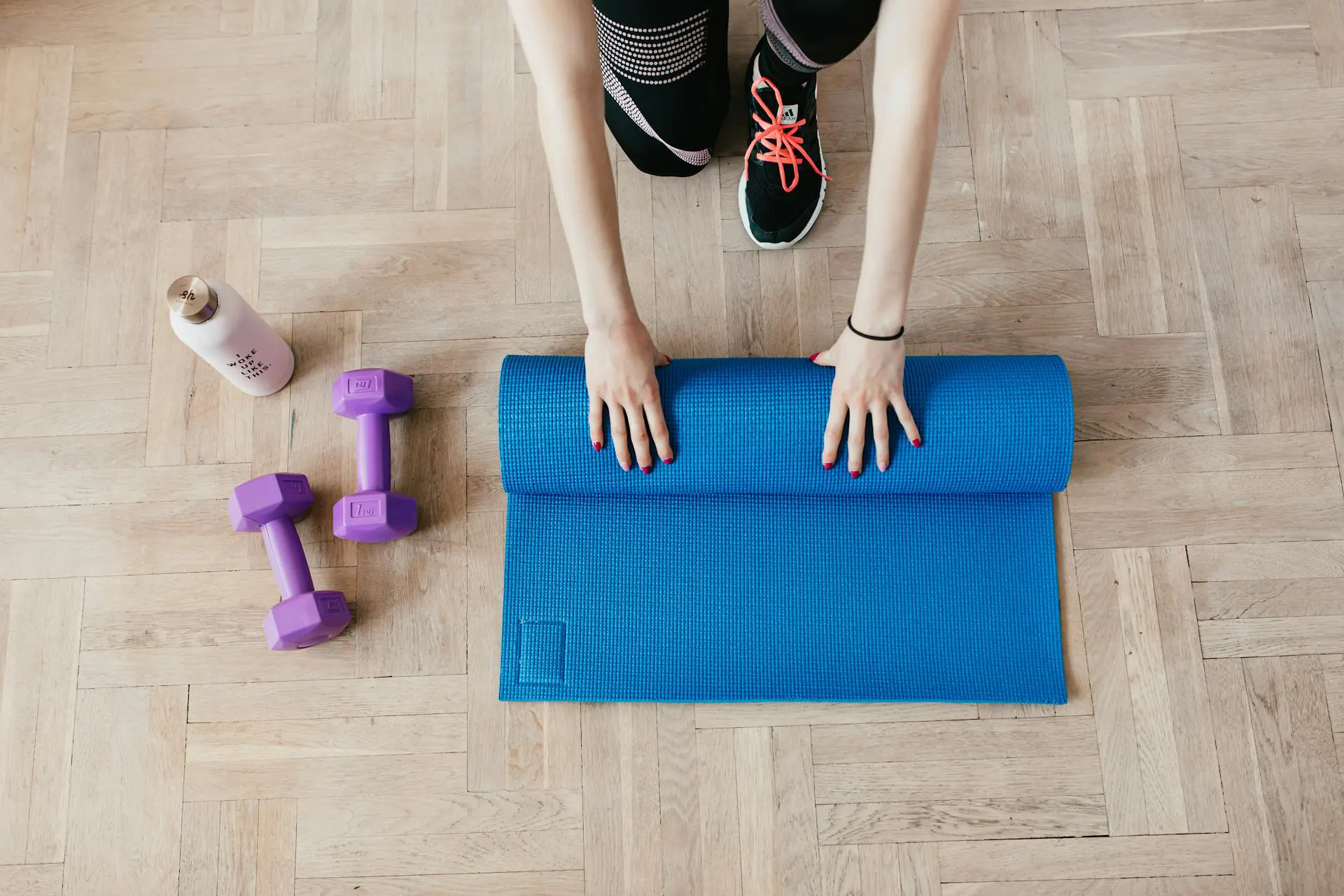 Nell'immagine c'è una ragazza chinata a terra che piega un materassino da yoga blu. Accanto a lei ci sono due pesetti viola e una borraccia